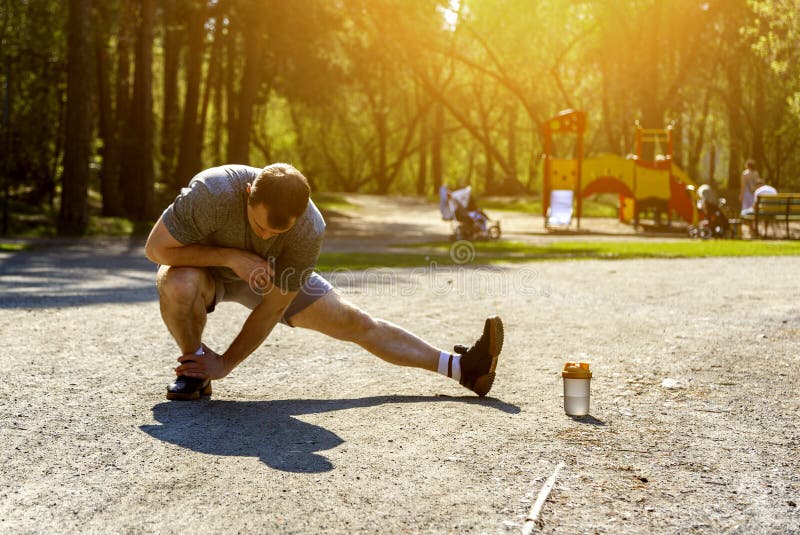 Healthy caucasian athlete stretching legs before run. Under rays of sunset. Athletic sportsman doing warm-up exercises. Prepared bottle with water. Playground royalty free stock images