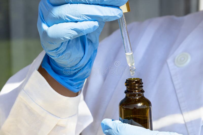 Woman hand in white coat and blue medical gloves holding pipette dropper with drop of natural oil above brown glass bottle royalty free stock images