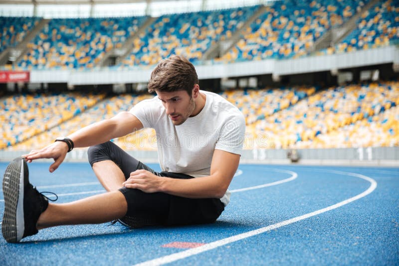 Young healthy sportsman stretching legs. While sitting on a racetrack at the stadium stock photography