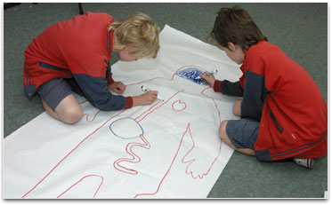 2 students are drawing onto an outline of a body on a large piece of paper.