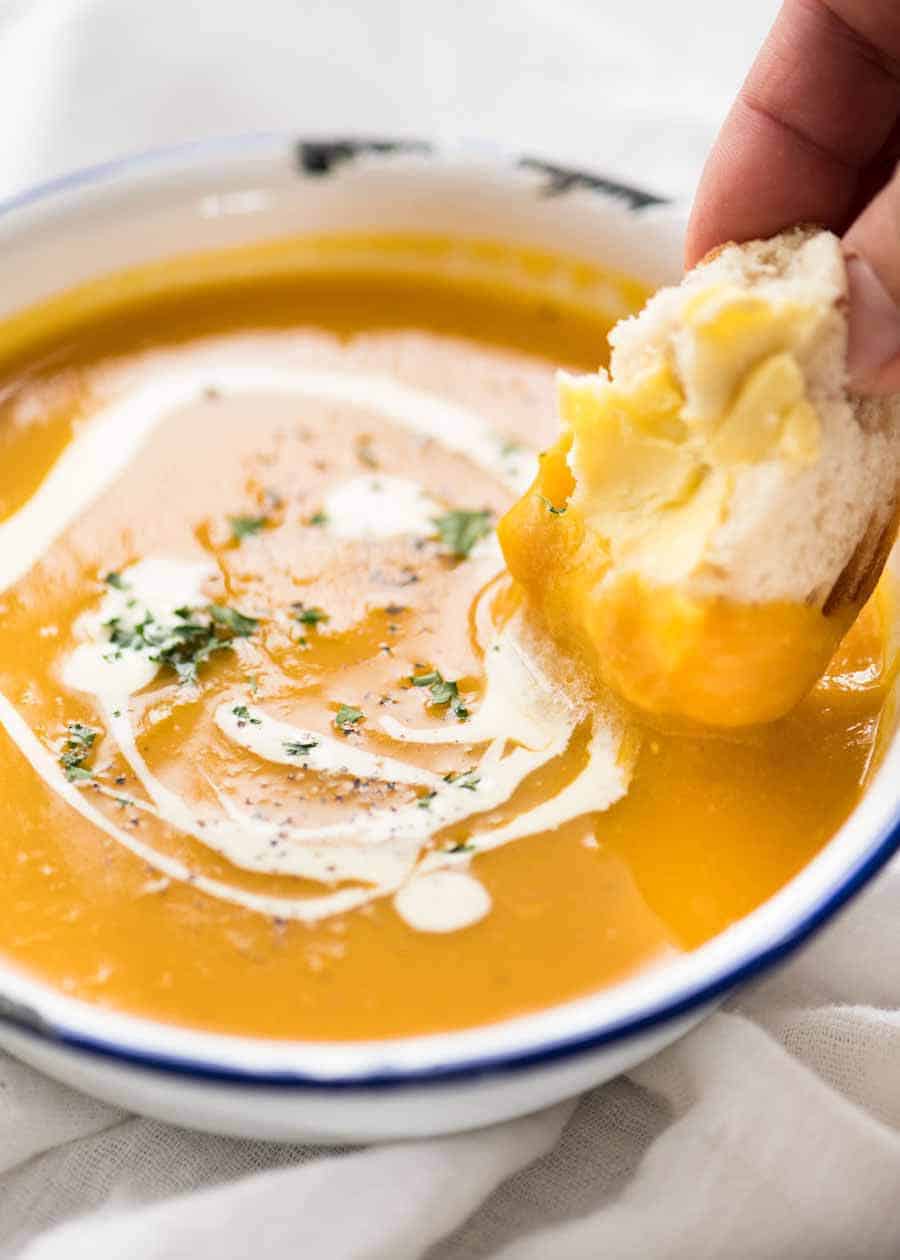 Close up of dunking crusty bread into thick and creamy pumpkin soup in a rustic white enamel bowl.